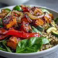 A bowl of warm salad featuring quinoa, roasted sweet potato, red bell pepper, and wilted spinach, drizzled with a golden warm vinaigrette and topped with feta and pumpkin seeds.